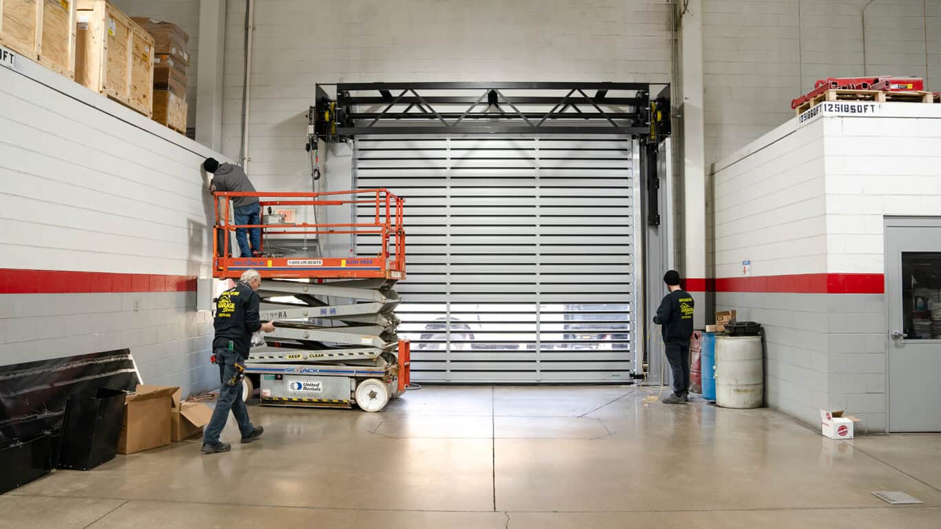 Workers installing a large garage door in an industrial setting with two people on a lift and one standing on the ground.