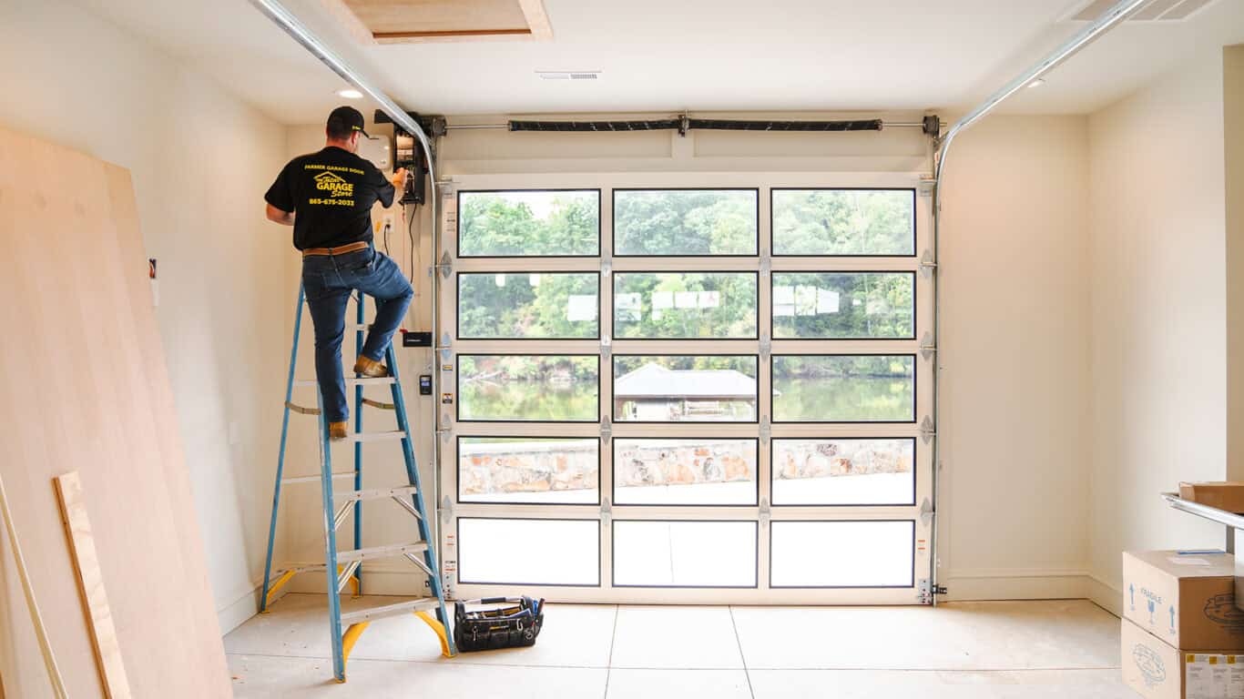 A technician on a ladder is carefully repairing a large glass-paneled residential garage door in a well-lit space, with trees visible outside.