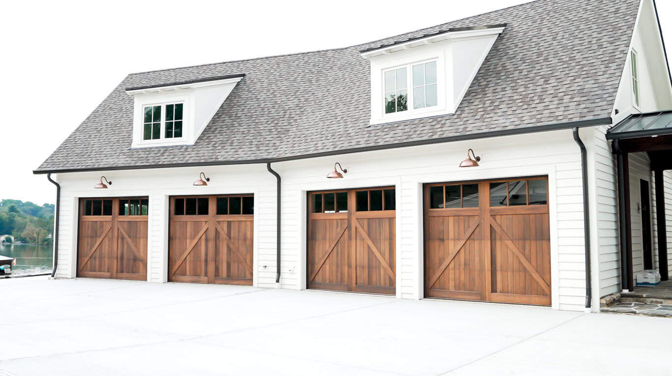 A four-car garage with wooden doors and a white exterior. The building has a shingled roof and a dormer window, set in a spacious driveway area.
