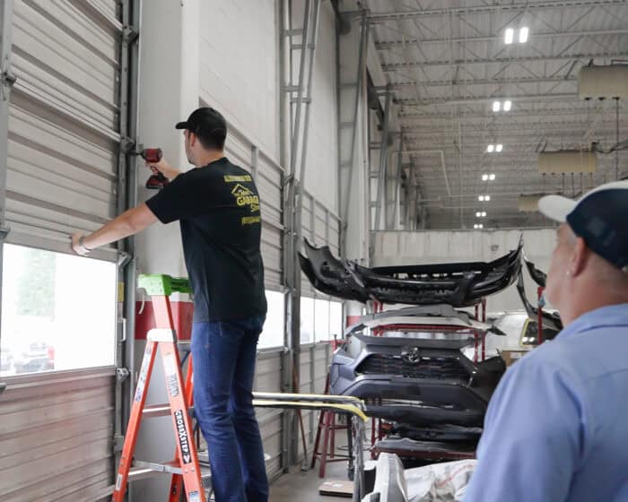Two men in a garage. One stands on a ladder fixing a garage door, while the other watches. Car parts and tools are visible in the background.