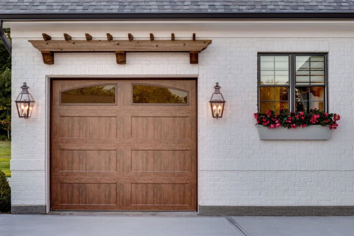 A wooden garage door with decorative windows is flanked by two lantern-style lights. A window box with red flowers and greenery is on the right wall.
