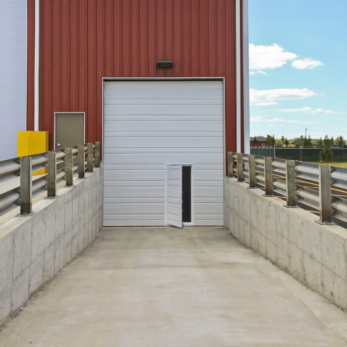 A warehouse loading dock features a large commercial overhead door in white, with a smaller open entryway within it. A concrete ramp, flanked by metal railings on both sides, provides easy access.