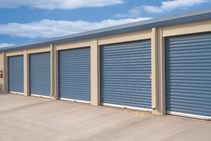 A row of closed blue roll-up doors lines the beige storage unit building under a cloudy sky, reflecting expert garage door installation.